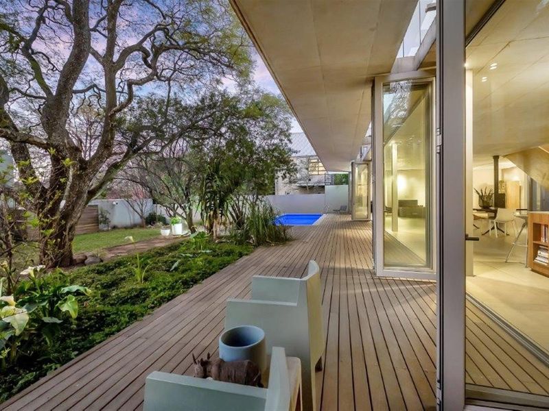 A modern covered timber deck overlooking a landscaped garden and swimming pool, seamlessly connecting to the interior living space through large glass sliding doors.