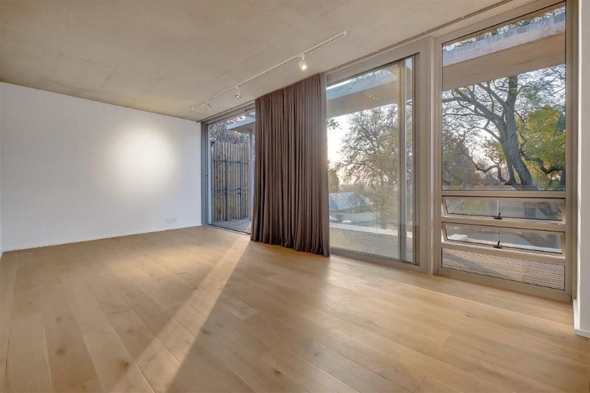 A bright, modern living room featuring light timber flooring, exposed concrete ceiling, and large sliding glass doors opening onto a private balcony.