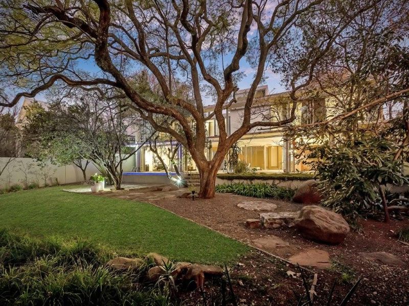 A beautifully landscaped rear garden featuring mature trees, a manicured lawn, and a swimming pool set against the rear facade of the house.
