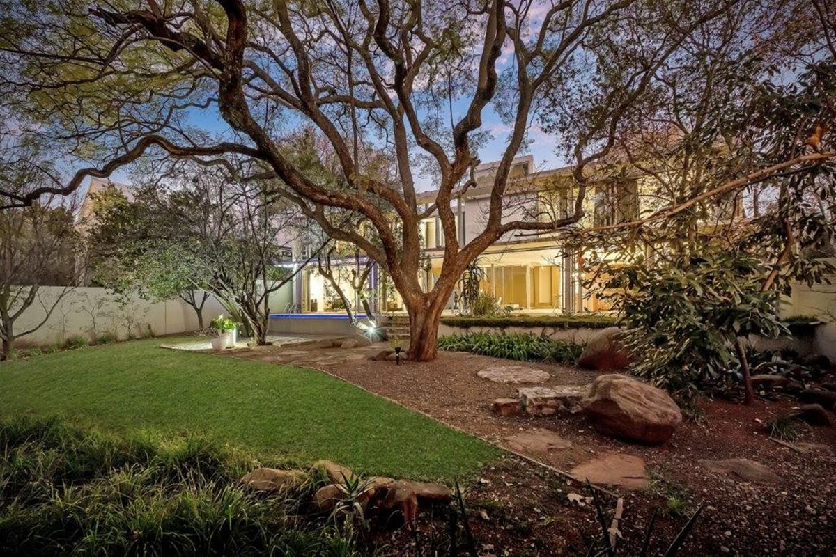 A beautifully landscaped rear garden featuring mature trees, a manicured lawn, and a swimming pool set against the rear facade of the house.
