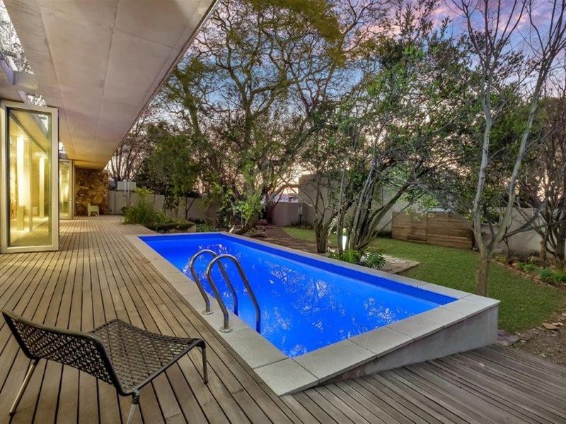 A well-maintained outdoor entertainment area featuring a rectangular swimming pool with stainless steel ladder, surrounded by a timber deck and landscaped garden.