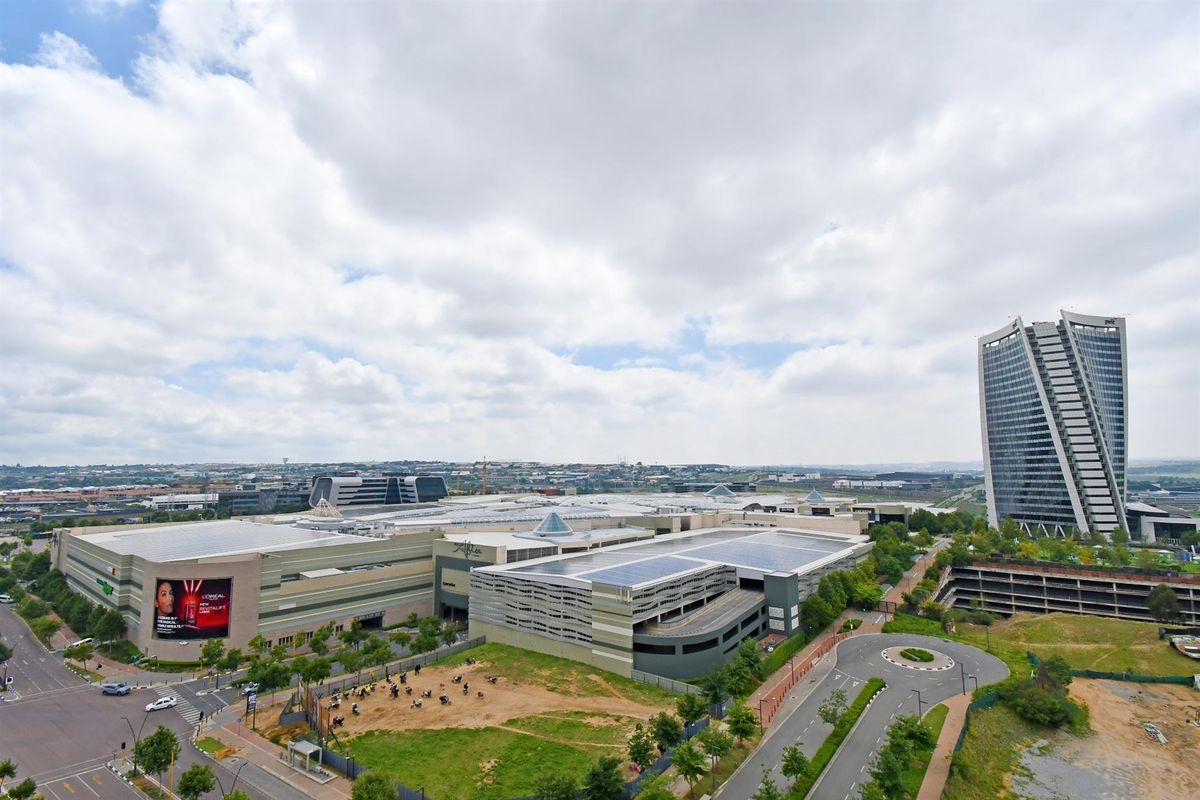 An aerial view of a large commercial complex featuring extensive parking facilities, solar panel installations, and surrounding urban road infrastructure.