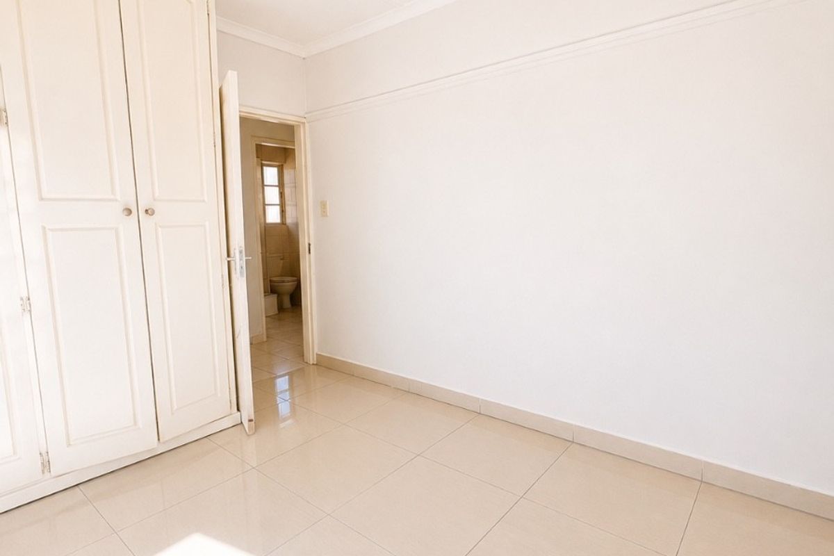 A bright, empty bedroom featuring neutral tiled flooring, built-in wardrobe storage, and a view into the adjacent bathroom.