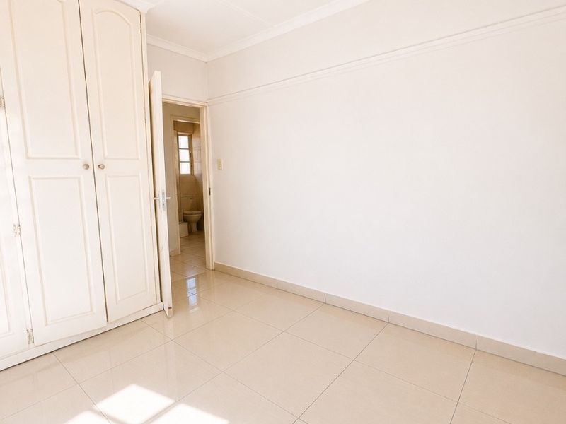 A bright, empty bedroom featuring neutral tiled flooring, built-in wardrobe storage, and a view into the adjacent bathroom.
