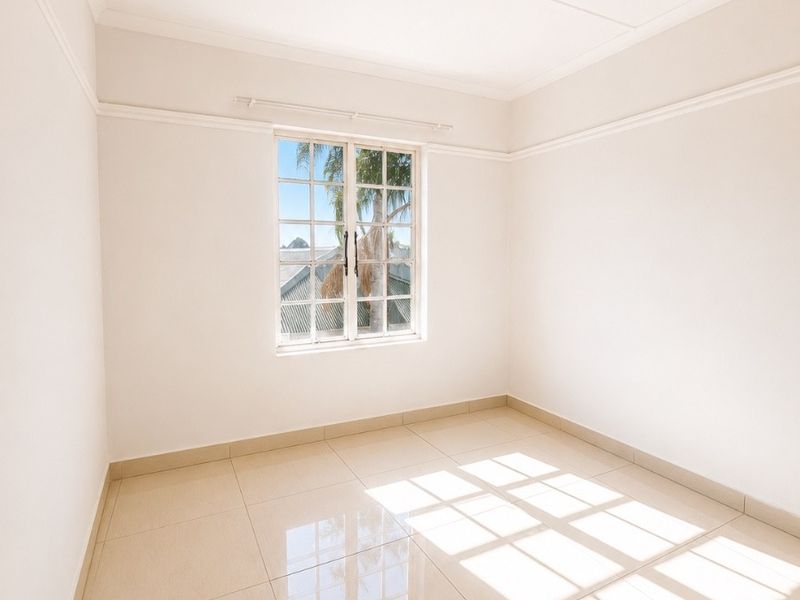 A bright, empty bedroom featuring neutral tiled flooring, crisp white walls, and a classic lattice window providing natural light.