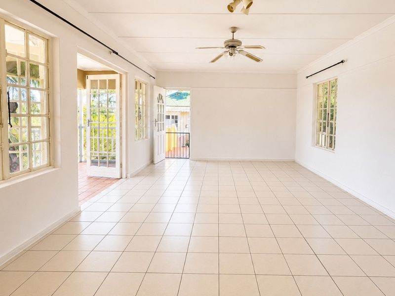 A spacious, empty living area featuring neutral tiled flooring, a ceiling fan, and multiple windows and doors providing natural light and access to the exterior.