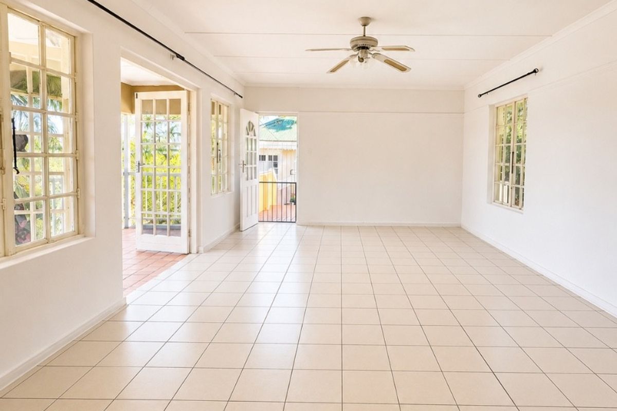 A spacious, empty living area featuring neutral tiled flooring, a ceiling fan, and multiple windows and doors providing natural light and access to the exterior.