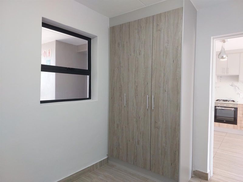 A clean, modern bedroom featuring built-in wood-look cupboards, tiled flooring, and a view through to the adjacent kitchen area.