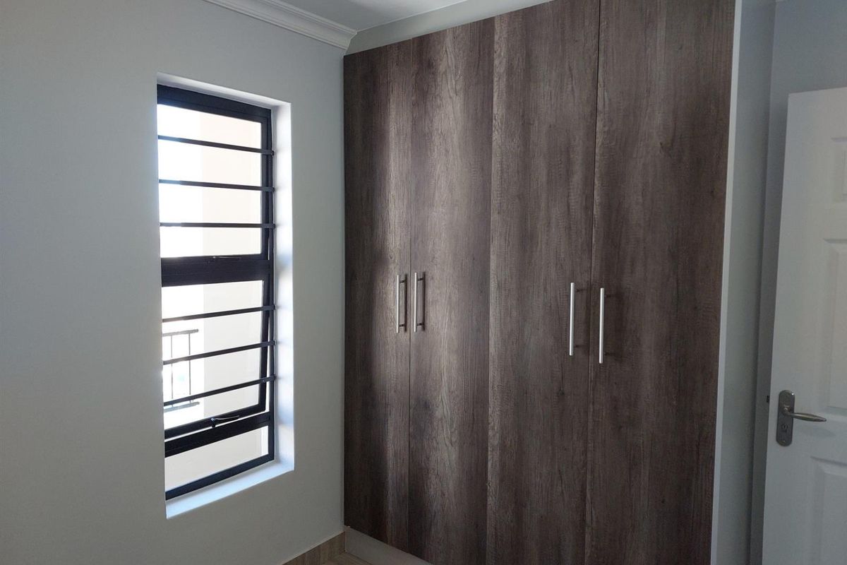 A clean, empty bedroom featuring modern dark wood-grain built-in cupboards, a window with security bars, and neutral wall finishes.