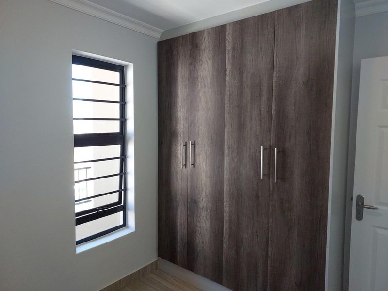 A clean, empty bedroom featuring modern dark wood-grain built-in cupboards, a window with security bars, and neutral wall finishes.