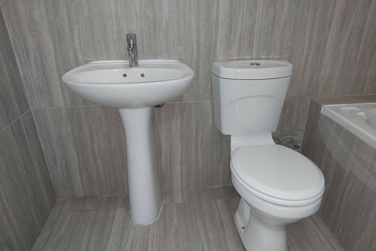 A functional bathroom featuring a pedestal basin, close-coupled toilet, and tiled finishes, with some visible damage to the wall tiling behind the toilet.