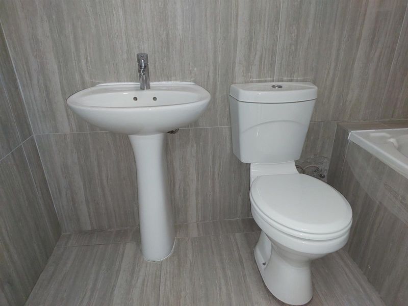 A functional bathroom featuring a pedestal basin, close-coupled toilet, and tiled finishes, with some visible damage to the wall tiling behind the toilet.
