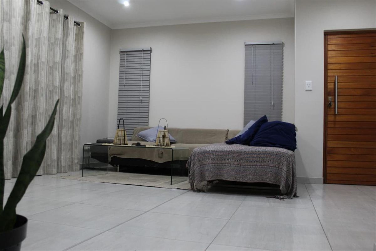 A well-lit living room featuring neutral tiled flooring, modern recessed downlights, and a solid timber entrance door with venetian blinds.