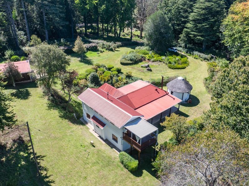 Aerial view of a residential property featuring a main house with a metal roof, a separate thatched outbuilding, and extensive landscaped garden grounds.