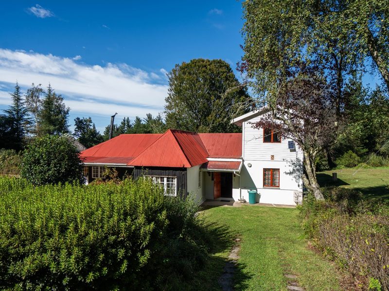 Rear view of a two-story house featuring a red corrugated metal roof, white timber siding, and a surrounding garden area.