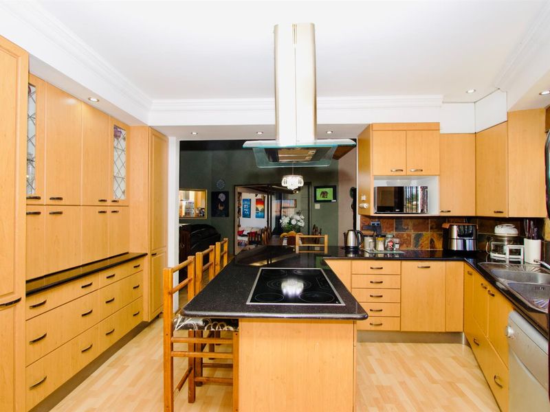 A functional kitchen featuring light wood cabinetry, black granite countertops, a central island with an integrated electric hob, and a stainless steel extractor fan.