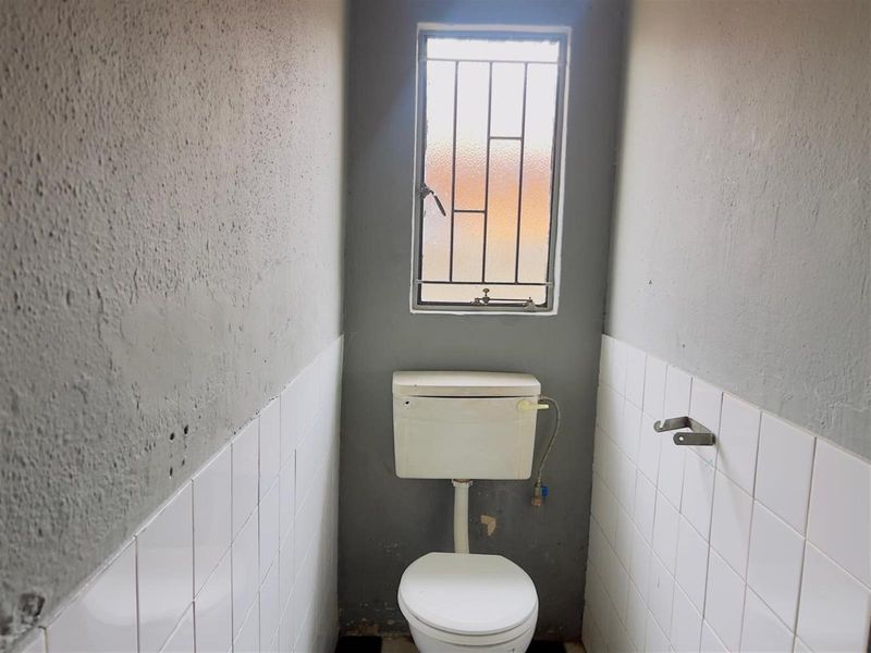 A small, basic bathroom featuring a toilet, security bars on the window, and partially tiled walls requiring cosmetic updates and finishing.