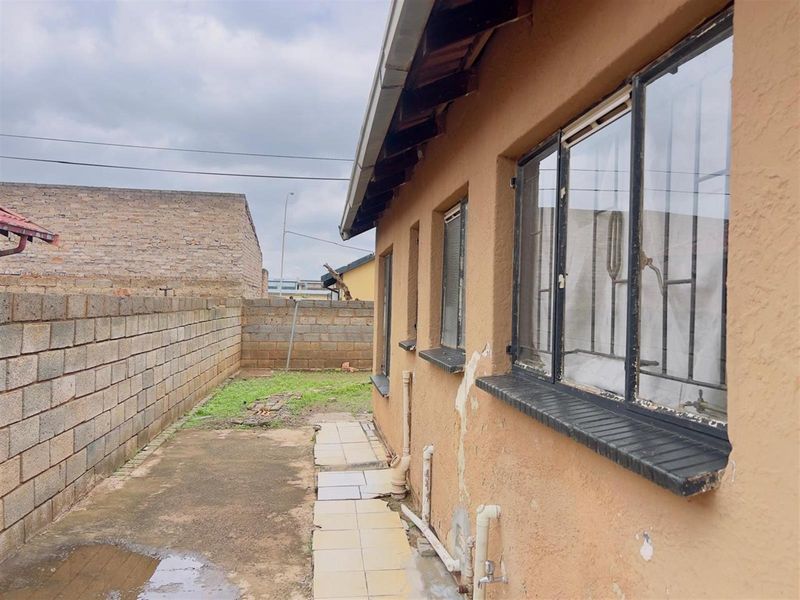 Rear exterior view of the property showing a side pathway, brick boundary wall, and the side facade with metal window frames and security bars.