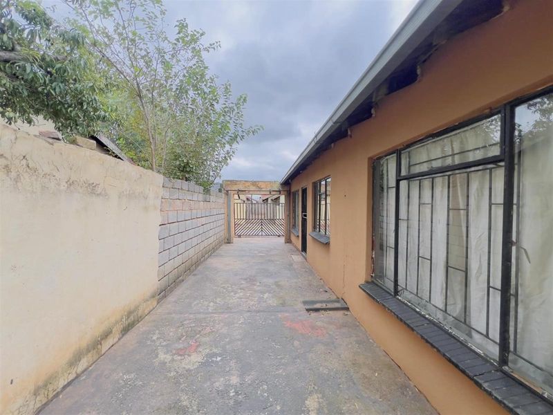 A side exterior view of the property featuring a concrete driveway, perimeter boundary wall, and metal security gate with window security bars.