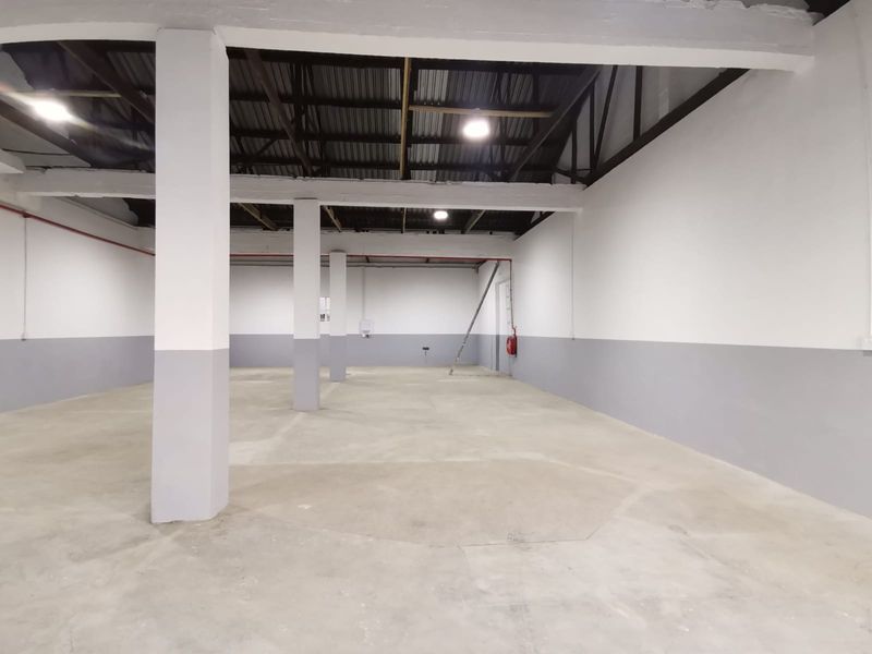 Spacious industrial warehouse area featuring high ceilings, exposed structural beams, concrete flooring, and functional overhead lighting.