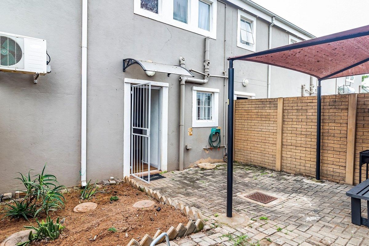Rear exterior view of the townhouse featuring a paved patio area, shade sail structure, security gate, and wall-mounted air conditioning unit.