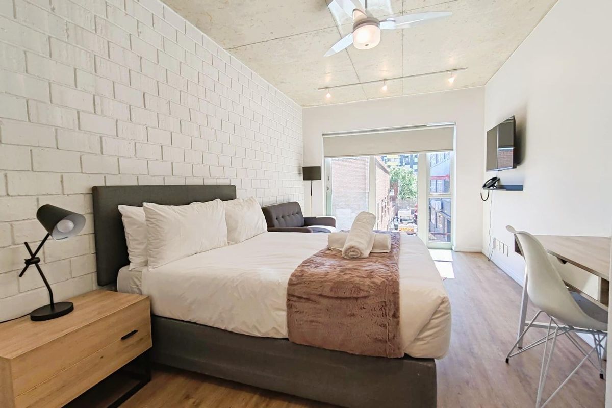 Modern bedroom featuring an exposed concrete ceiling, white painted brick feature wall, wood-look flooring, and a glass door leading to a balcony.