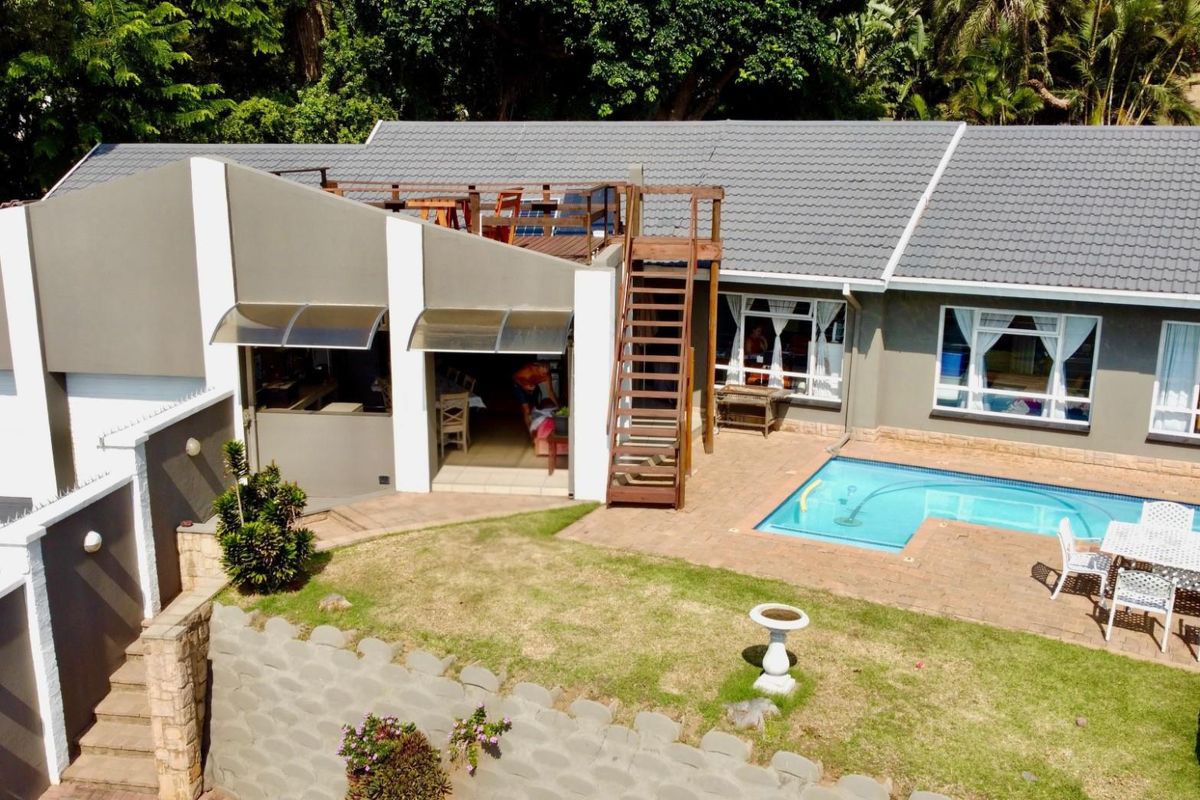 Aerial view of the property featuring a swimming pool, paved patio area, rooftop deck with wooden staircase, and a well-maintained garden.