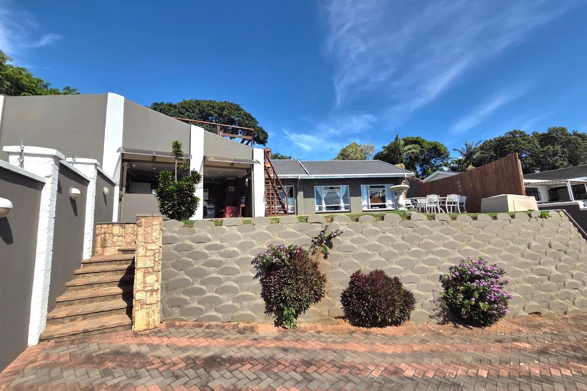 Rear view of the property featuring a tiered retaining wall, stone staircase, paved driveway, and an elevated timber deck area.