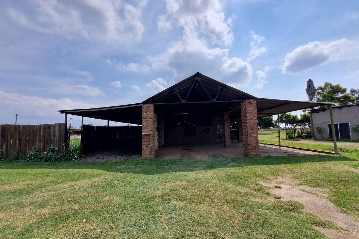 A large open-sided farm structure featuring exposed brick pillars and a metal roof, set within a spacious grassed area.