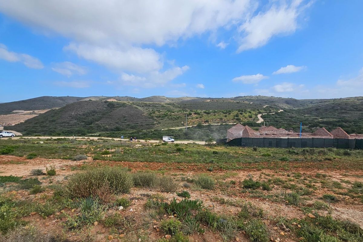 A wide-angle view of a vacant residential plot featuring natural landscape, rolling hills in the background, and ongoing construction activity on the adjacent site.