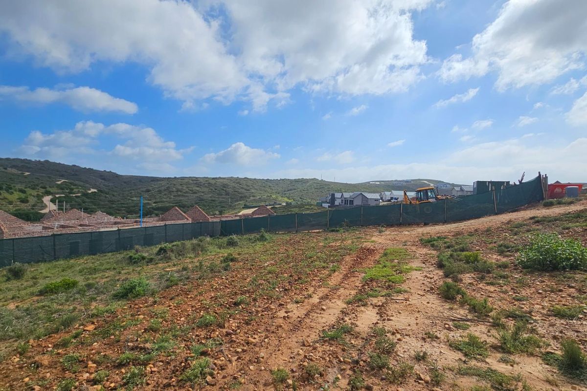 A view of a vacant residential plot featuring natural terrain, perimeter fencing, and surrounding development in a developing estate.