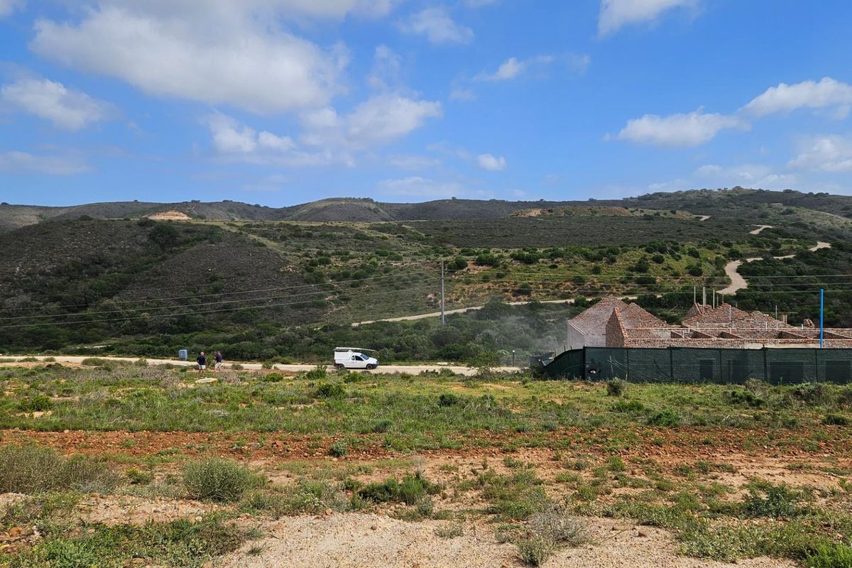 A wide-angle view of a vacant residential plot featuring natural landscape and an ongoing construction project in the background.