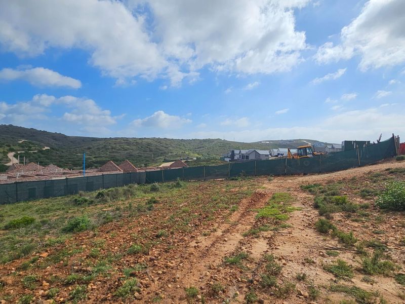 A view of a vacant residential plot featuring natural terrain, perimeter fencing, and surrounding development in a developing estate.