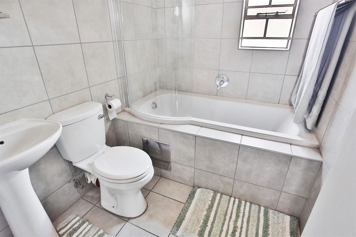A clean, functional bathroom featuring a pedestal basin, toilet, and a bathtub with a glass shower screen, finished with neutral wall and floor tiles.