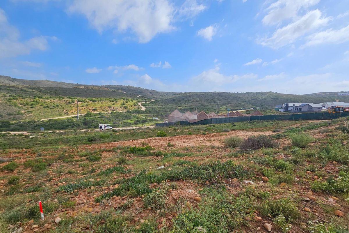 A wide-angle view of a vacant residential plot featuring natural landscape, rolling hills, and surrounding development in the distance.