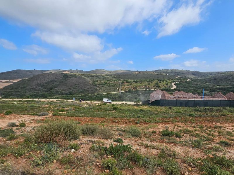 A wide-angle view of a vacant residential plot featuring natural landscape, rolling hills in the background, and ongoing construction activity on the adjacent site.