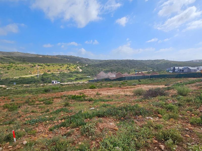 A wide-angle view of a vacant residential plot featuring natural landscape, rolling hills, and surrounding development in the distance.