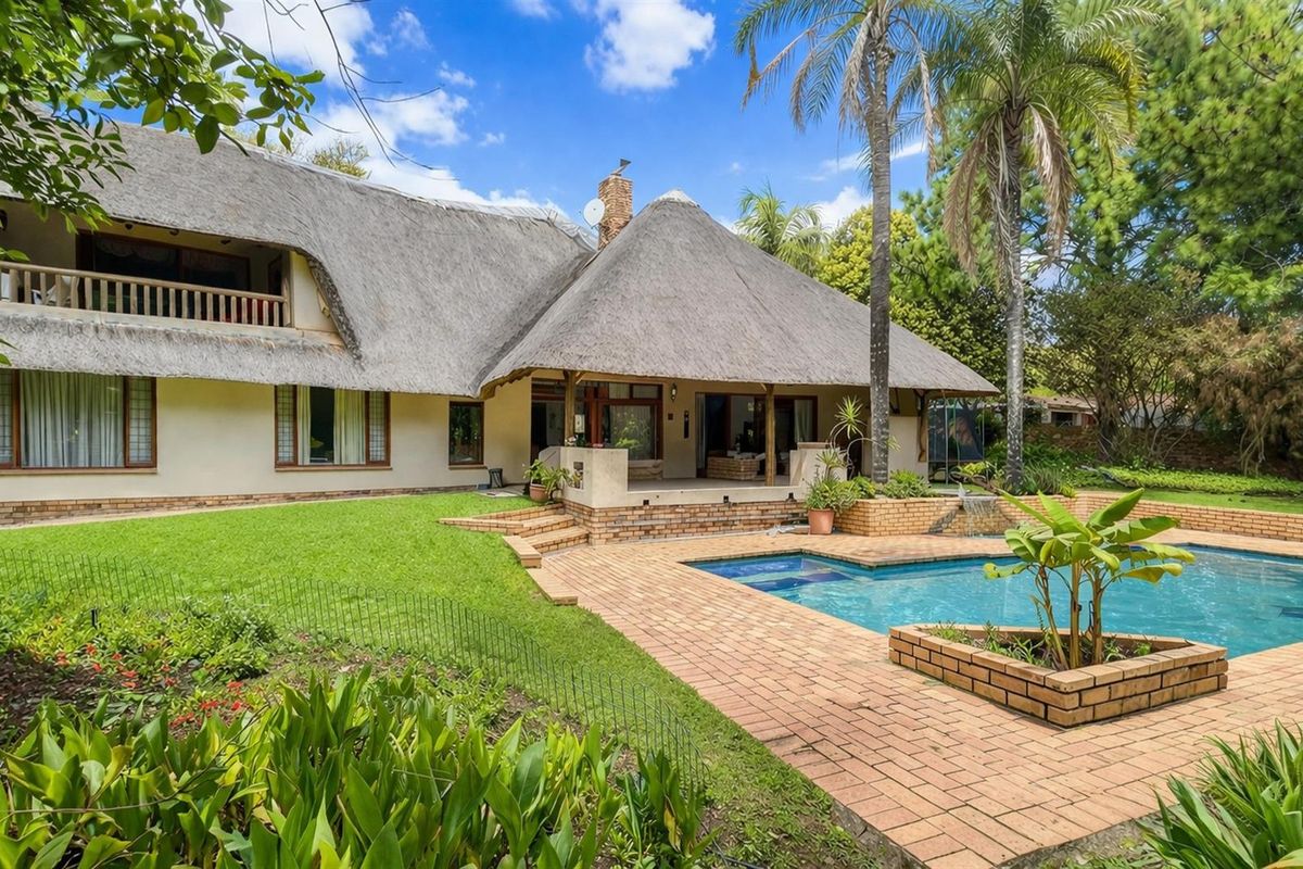 Rear view of a thatched-roof house featuring a swimming pool, brick-paved patio area, and established garden surroundings.