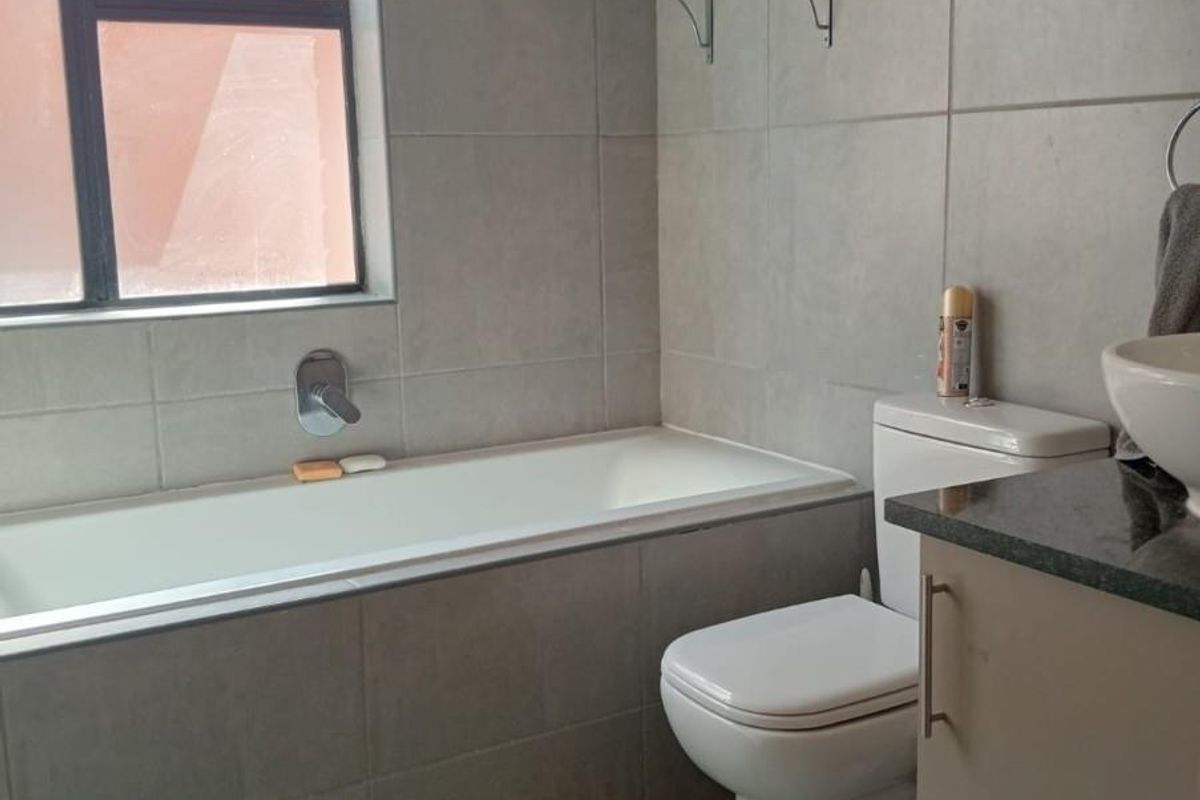 A modern bathroom featuring grey wall tiles, a built-in bathtub with chrome fixtures, a white toilet, and a vanity with a stone countertop.
