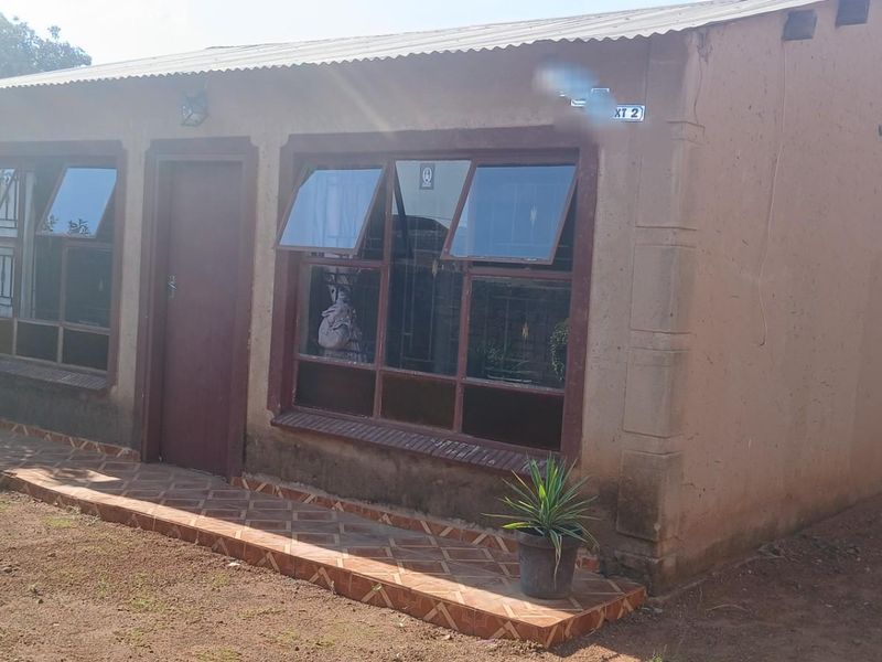 A modest single-story dwelling featuring a corrugated metal roof, brown-framed windows, and a tiled entrance walkway set on a dirt plot.