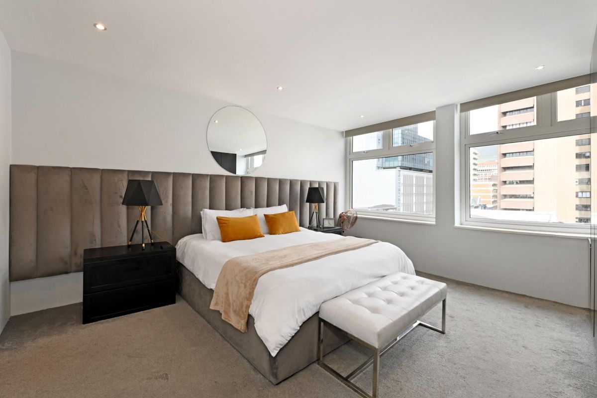 A well-lit bedroom featuring a large upholstered headboard, neutral carpeting, and expansive windows offering an urban city view.