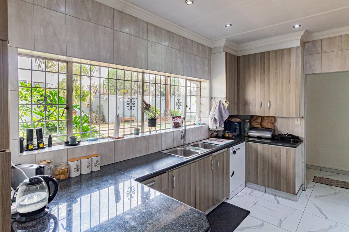 Modern kitchen featuring granite countertops, wood-look cabinetry, double stainless steel sink, and large windows with security bars overlooking the garden.