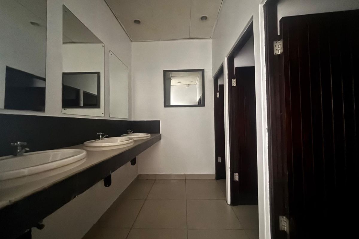 A functional commercial bathroom featuring a multi-basin vanity, tiled flooring, and multiple private cubicles with dark wood-finish doors.
