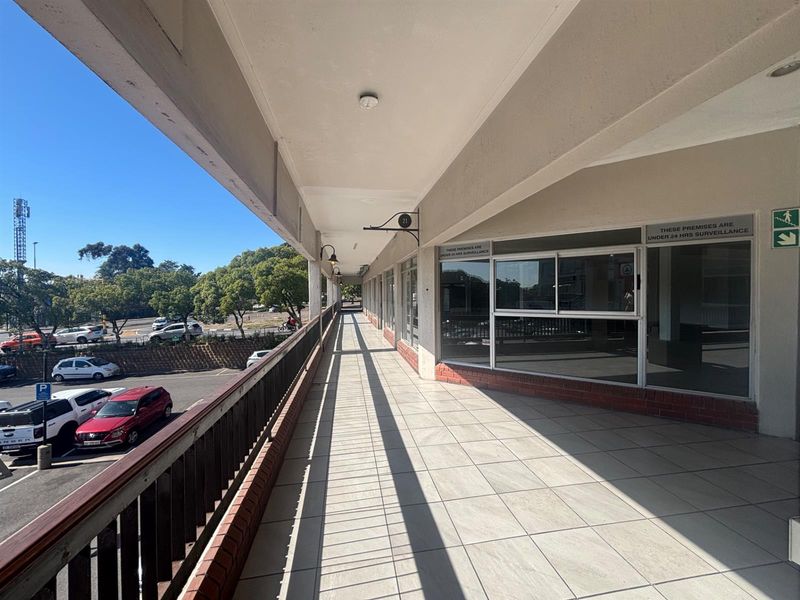 A covered exterior walkway or balcony featuring tiled flooring, metal railings, and large glass storefront windows overlooking a parking area.