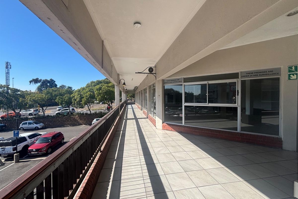 A covered exterior walkway or balcony featuring tiled flooring, metal railings, and large glass storefront windows overlooking a parking area.