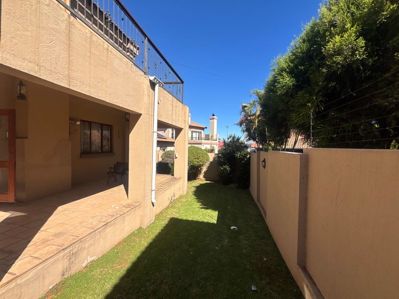 A rear exterior view featuring a covered patio with tiled flooring, a balcony above, and a small garden area enclosed by a boundary wall.