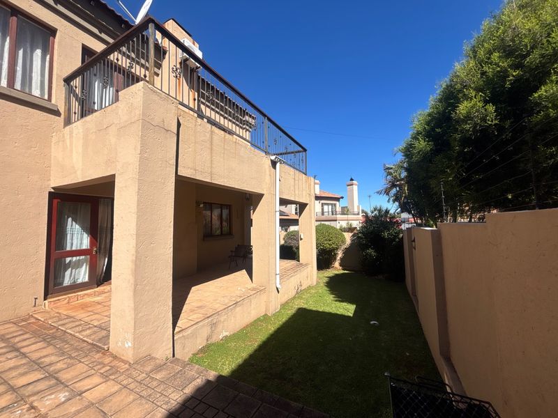 Rear view of the property featuring a covered patio, an upper-level balcony with metal railings, a small lawn, and perimeter walling with electric fencing.