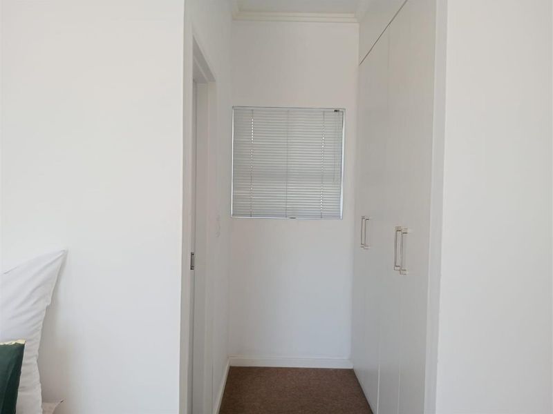 A clean bedroom hallway featuring built-in white cupboards, carpeted flooring, and a window fitted with venetian blinds.