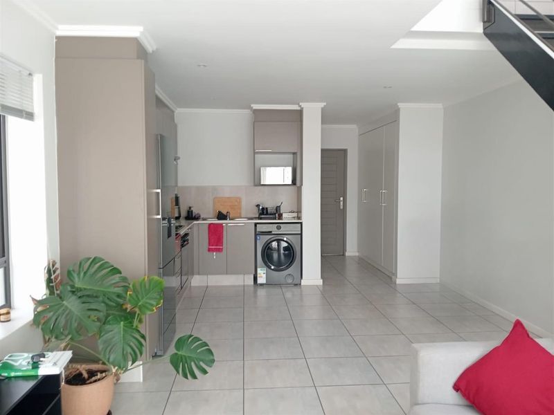 Modern open-plan living area featuring a compact kitchen with built-in cabinetry, tiled flooring, and integrated laundry facilities, leading to a staircase.