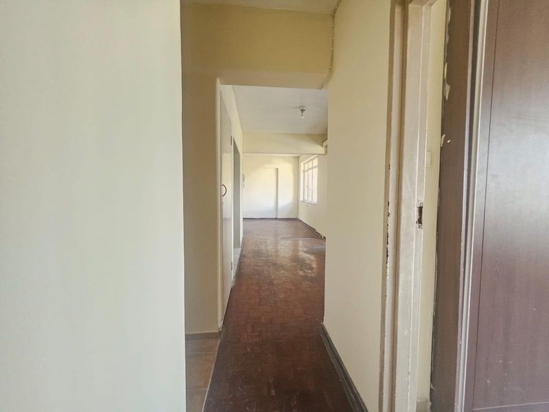 An empty hallway featuring worn parquet flooring and neutral walls, leading into an open living area with natural light.